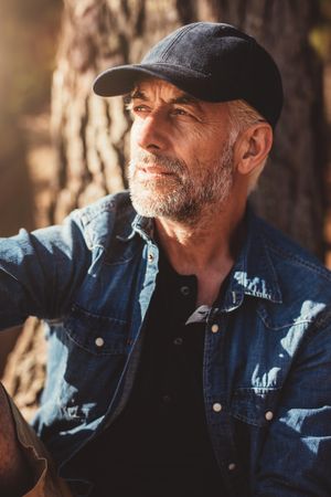Close up portrait of mature man wearing cap looking away while sitting by a tree