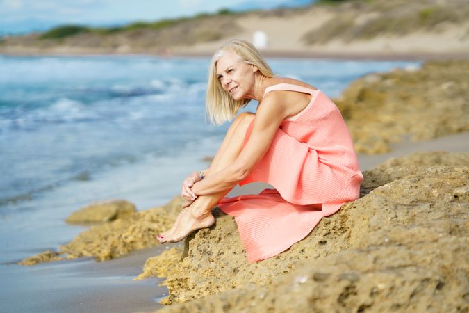 Older female with grey hair sitting on coastal rocks enjoying the view