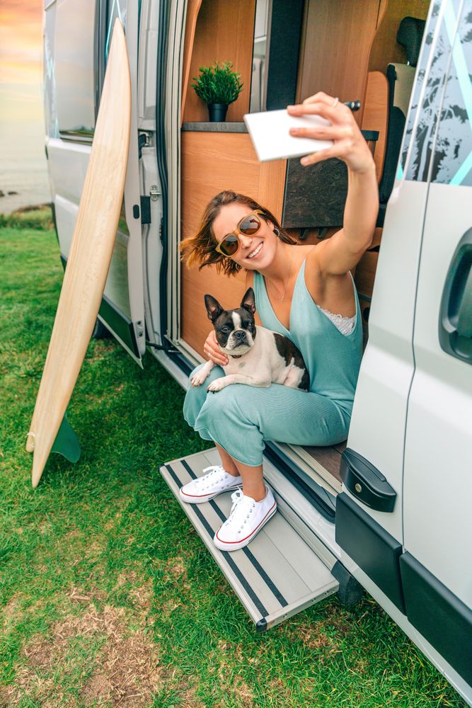 Smiling female sitting stoop of motorhome taking selfie with cute dog, vertical