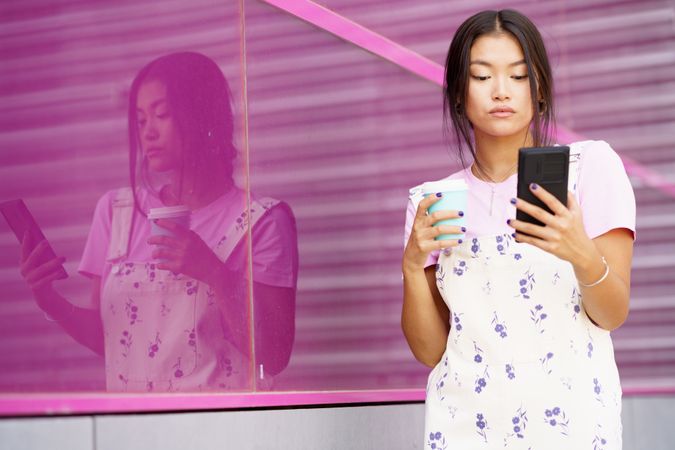 Female in floral overalls checking her smartphone with a takeaway coffee