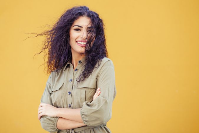 Female in army green jumpsuit smiling in front of mustard wall with arms folded, copy space