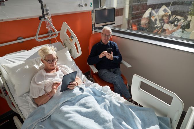 Mature man sitting near sick wife lying on couch and using table