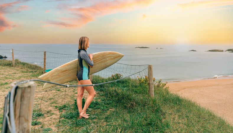 Woman standing on cliff overlooking beach holding a surfboard