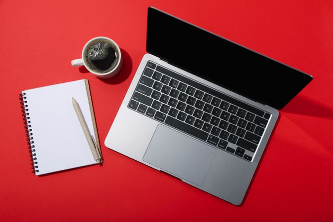 Laptop with blank screen on red table with coffee and notebook