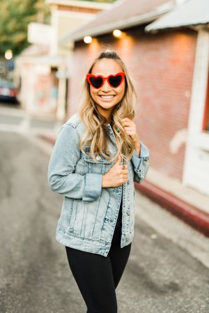 Woman with red framed sunglasses in denim jacket standing on sidewalk
