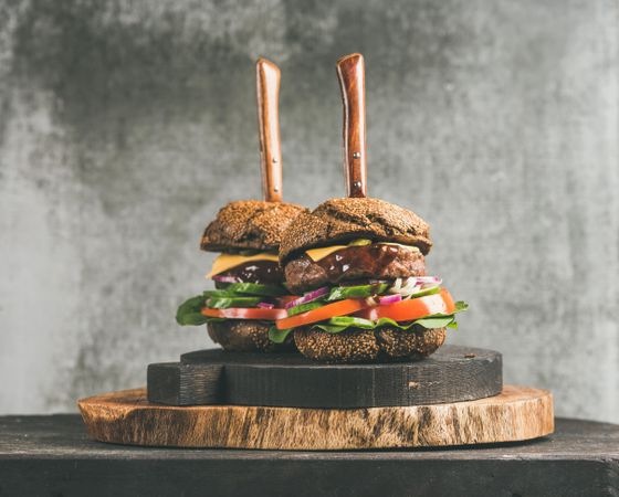 Two cheeseburgers skewered with knives, with fresh vegetables, on wooden board