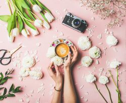 Coffee, camera, white tulips artfully arranged on pink background, with hands on coffee cup