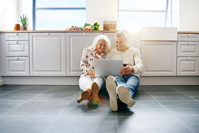 Grey haired couple watching laptop while sitting on kitchen floor