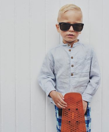 Young serious blond boy leaning against a wall with red skateboard