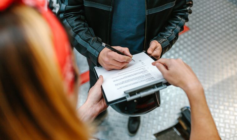 Female mechanic holding clipboard while biker man signing insurance policy on workshop