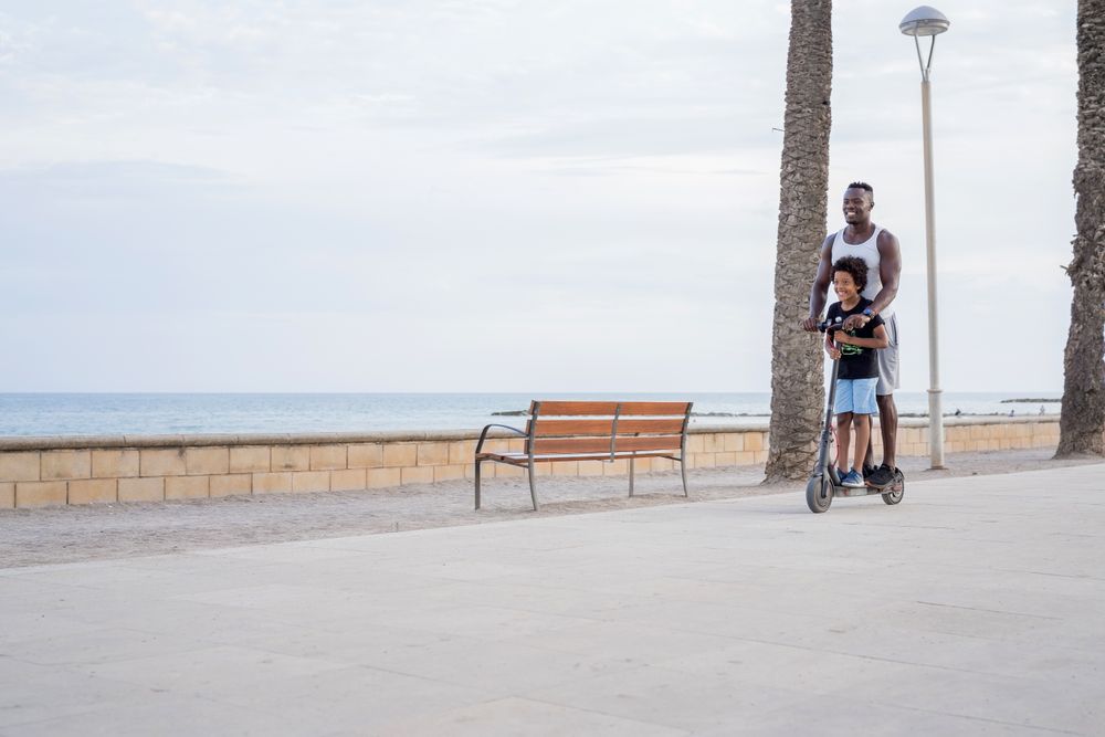 Cheerful Black father with kid riding scooter on promenade
