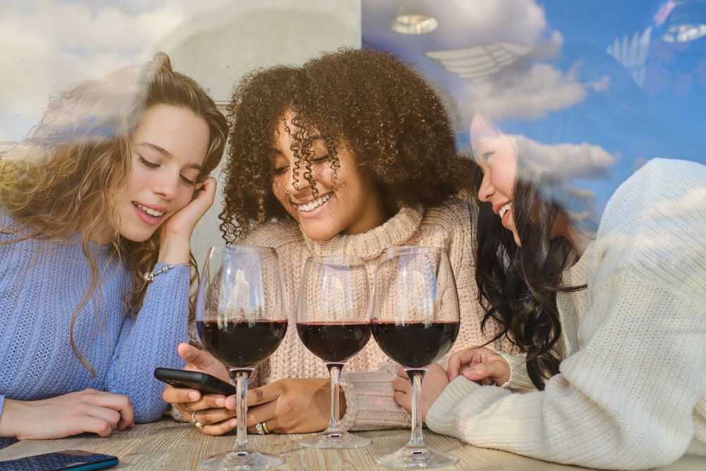 Smiling multiethnic women with wine looking at phone screen