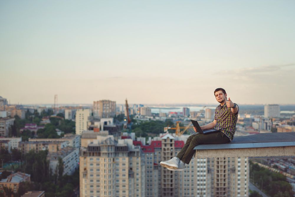 Side view of man perched on roof giving thumbs up with with laptop on lap