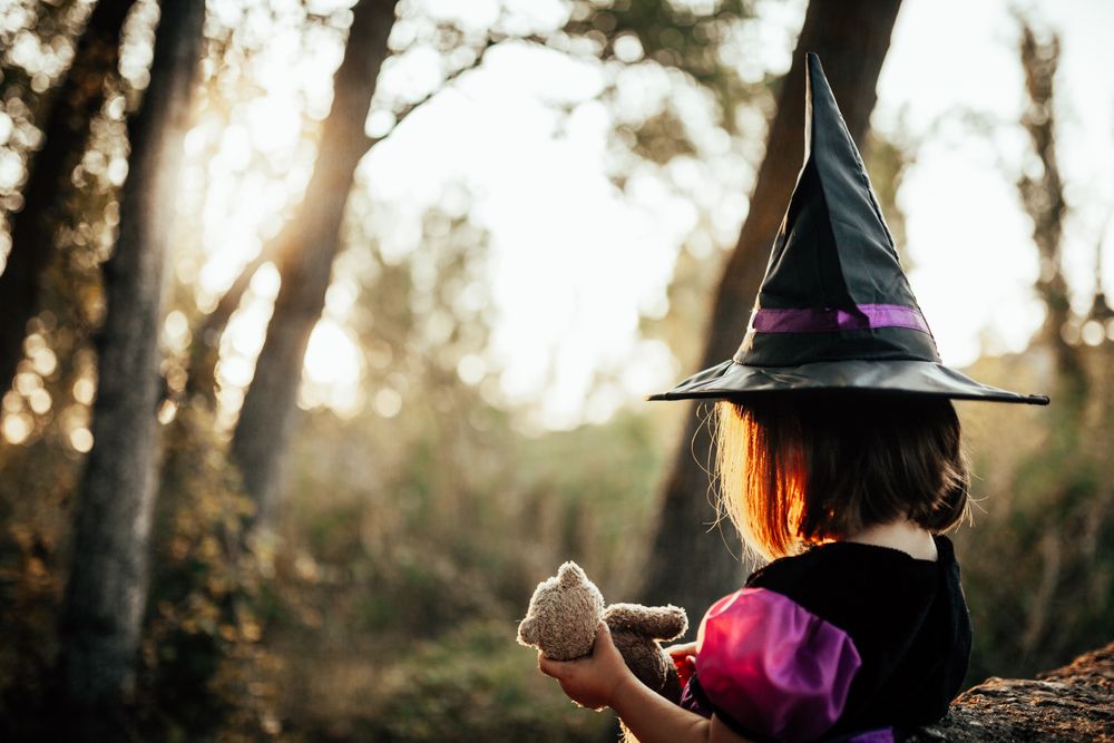 Girl in witch costume holding her teddy bear in the forest