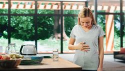 Young Adult Woman with Blond Hair Wearing Gray V-Neck T-Shirt, Rubbing Her Stomach After Food.