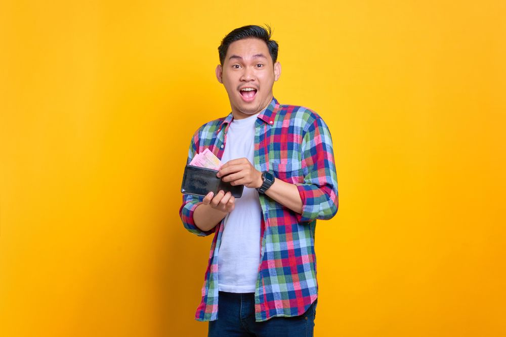 Excited Asian man smiling while taking cash out of his wallet