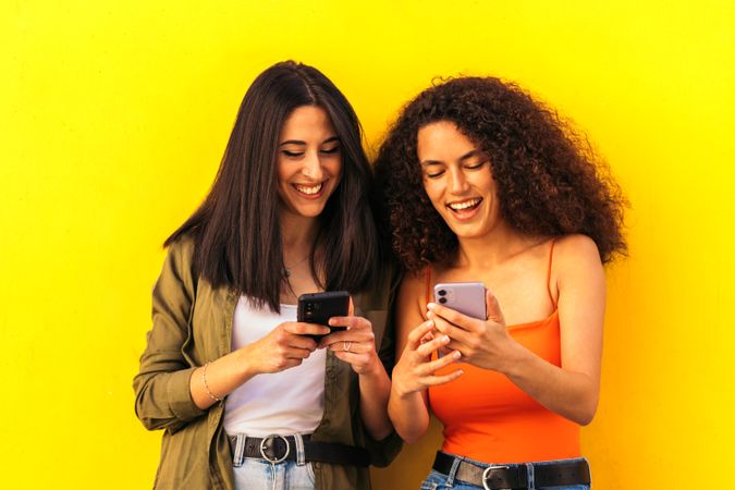 afro latina and white women watching mobile phone while leaning on a yellow wall