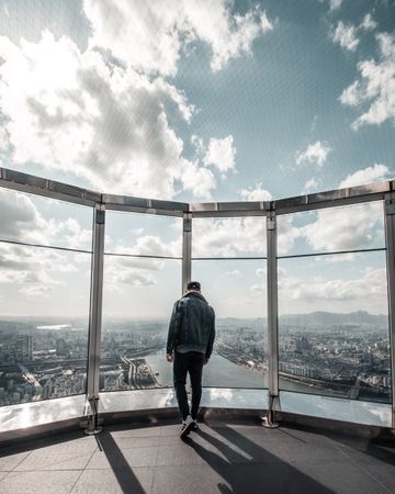 Back view of a man in denim jacket standing on a rooftop of skyscraper looking down the city