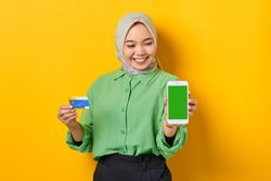 Happy Muslim woman in headscarf and green blouse holding credit card and smart phone screen mock up