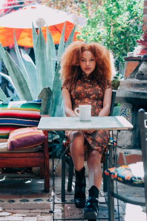 Young Black woman sitting at table outside with plants behind her