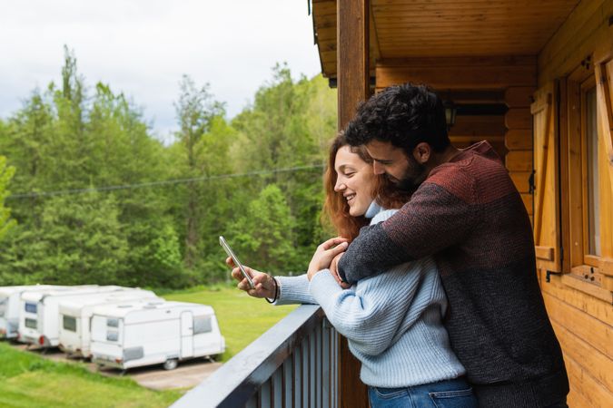 Cheerful traveling couple embracing and using smartphone on terrace of house