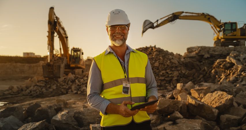Successful Bearded Civil Engineer Wearing Protective Goggles And Smiling At Camera On Construction Site on a Sunny Day While Heavy Machinery Working. Man Holding Tablet, Wearing Hard Hat, Safety Vest.