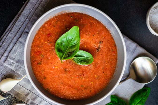 Top view of grey bowl of gazpacho soup with basil leaves and garlic on napkin