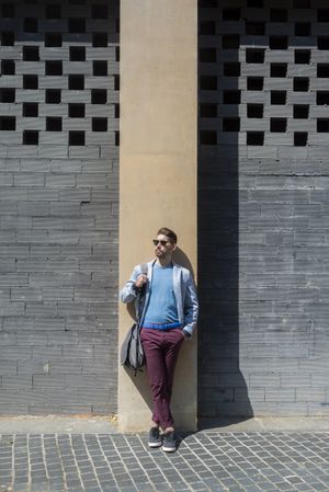 Bearded man wearing casual clothes while leaning on a building wall outside with bag