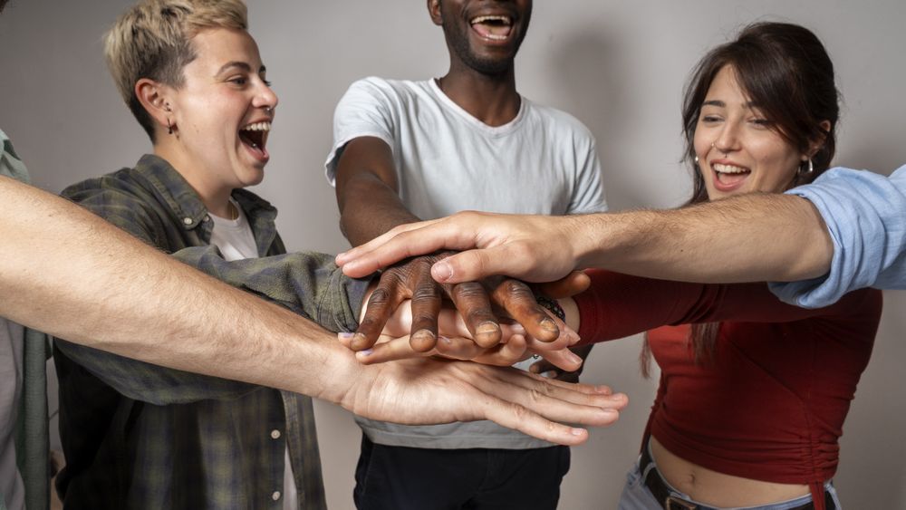 Team of diverse young adults joining hands in teamwork gesture, laughing and smiling together