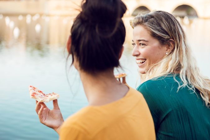 Smiling friends with slices of pizza sitting on Italian embankment