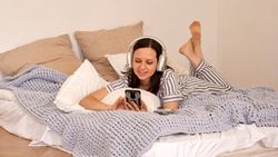 Woman lying in bed with headphones, using smartphone.