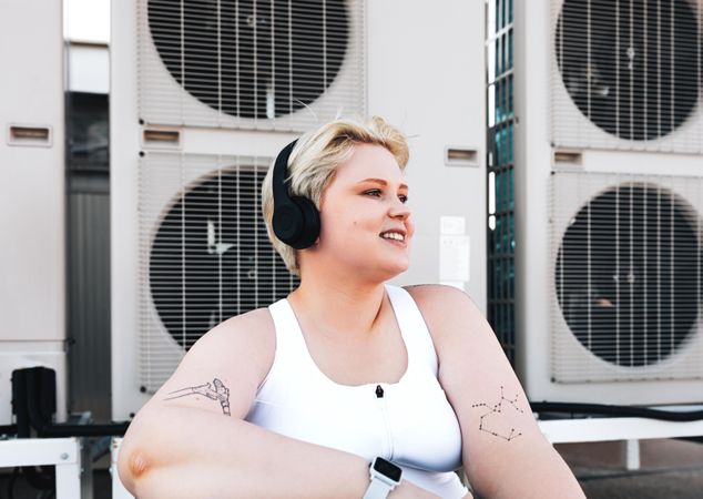 Woman with tattoos, smart watch and headphones smiling in front of fans on rooftop