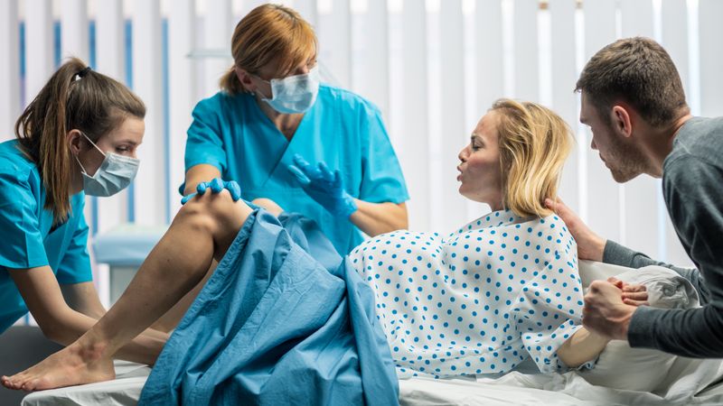 In the Hospital Woman in Labor Pushes to Give Birth, Obstetricians Assisting, Spouse Holds Her Hand