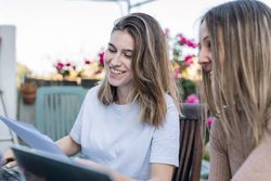 Two smiling friends comparing their notes and chatting about what was written on the laptop.