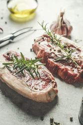 Close up of raw beef steak with green pepper and rosemary at grey kitchen table with butcher fork and olive oil. Preparing fresh meat for barbecue. Top view.