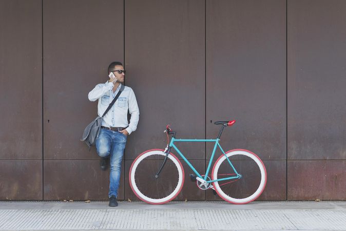 Male in sunglasses standing with red and green bicycle leaning on wall and speaking on phone