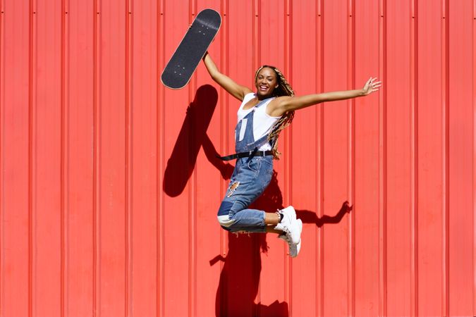 Woman smiling and jumping with arms up in front of a red wall with skateboard