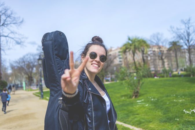 Side view of a young smiling hipster woman walking in a park in sunny day while carrying a guitar on back and gesturing victory sign