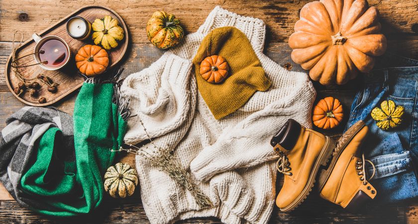 Flat-lay of beige knitted sweater, jeans, green woolen scarf, hat, yellow boots, pumpkins, candle