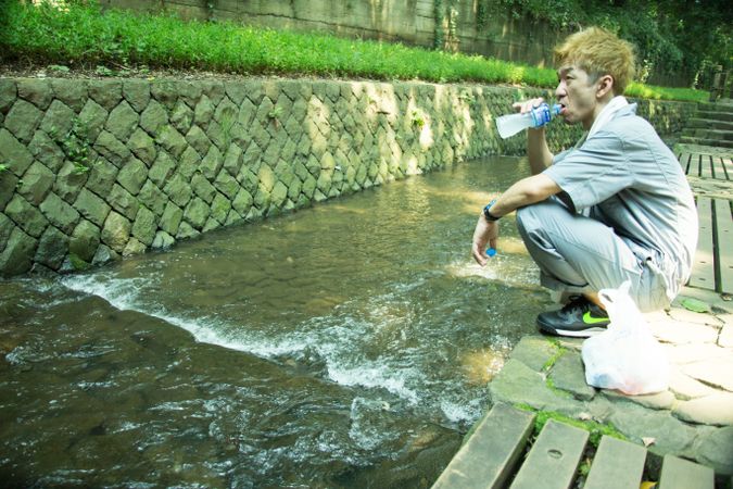 Man in work suit drinking water and crouching beside stream river