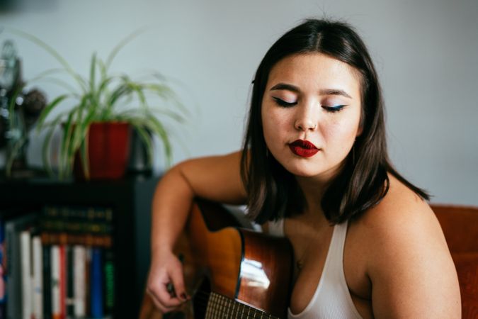 Horizontal shot of young woman sitting inside home playing guitar