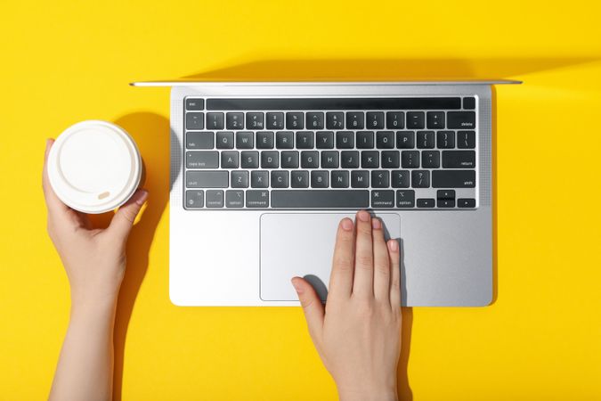 Top view of person using laptop with to go cup on yellow desk