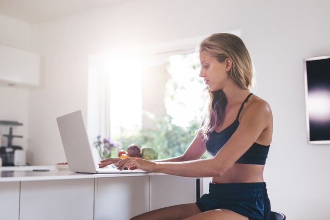 Side view of beautiful young woman sitting by kitchen counter and using laptop