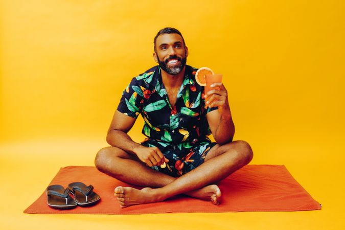 Black male sitting on towel and looking up while holding up tropical drink