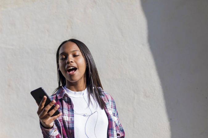 Female standing in the sun in front of wall singing along with music on phone