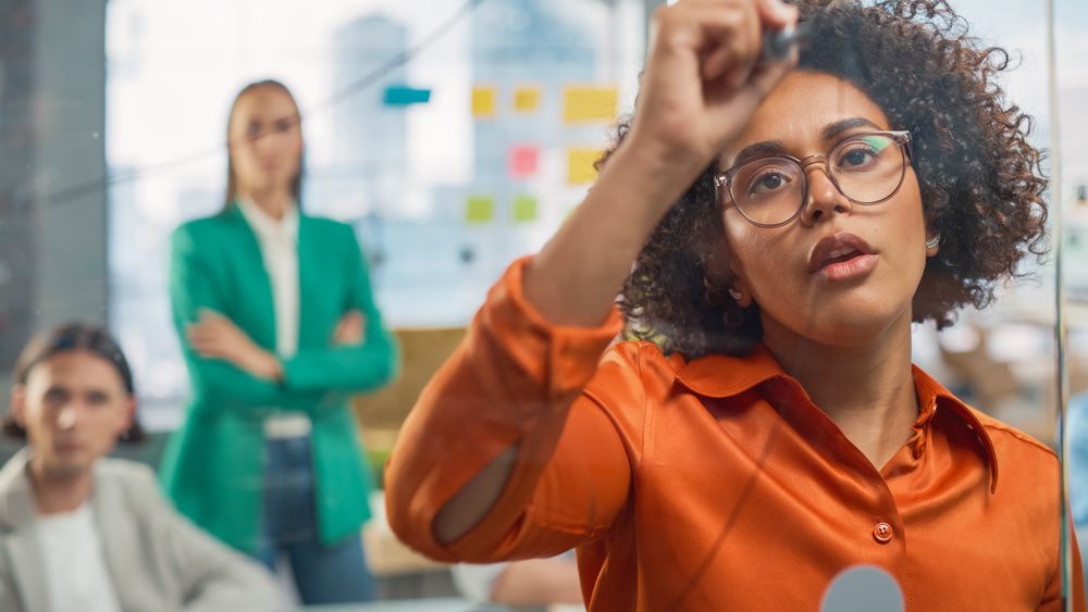 Portrait of Black Female Manager Writing on Glass Board and Brainstorming with Team in a Stylish Meeting Room in Creative Office.