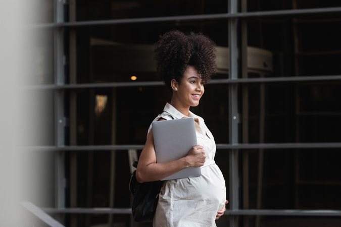 Smiling Black woman with laptop coming out of office building