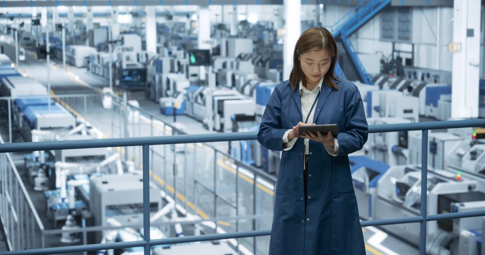 Japanese Factory Manager Working on a Tablet at a Futuristic Industrial Manufacture with Autonomous AI Robots. Specialist Monitoring All the Data on a Device, Checking Reports and Planning Research