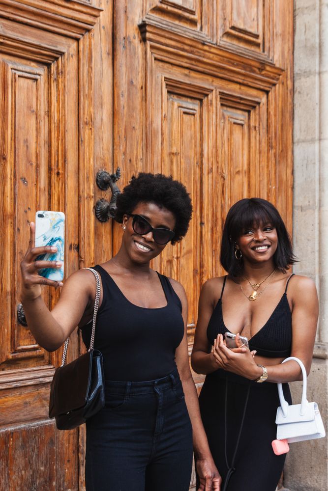Black females taking selfie with smartphones while standing on street