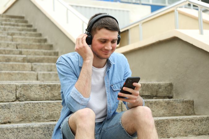 Young man in headphones sits on the steps and looking in phone